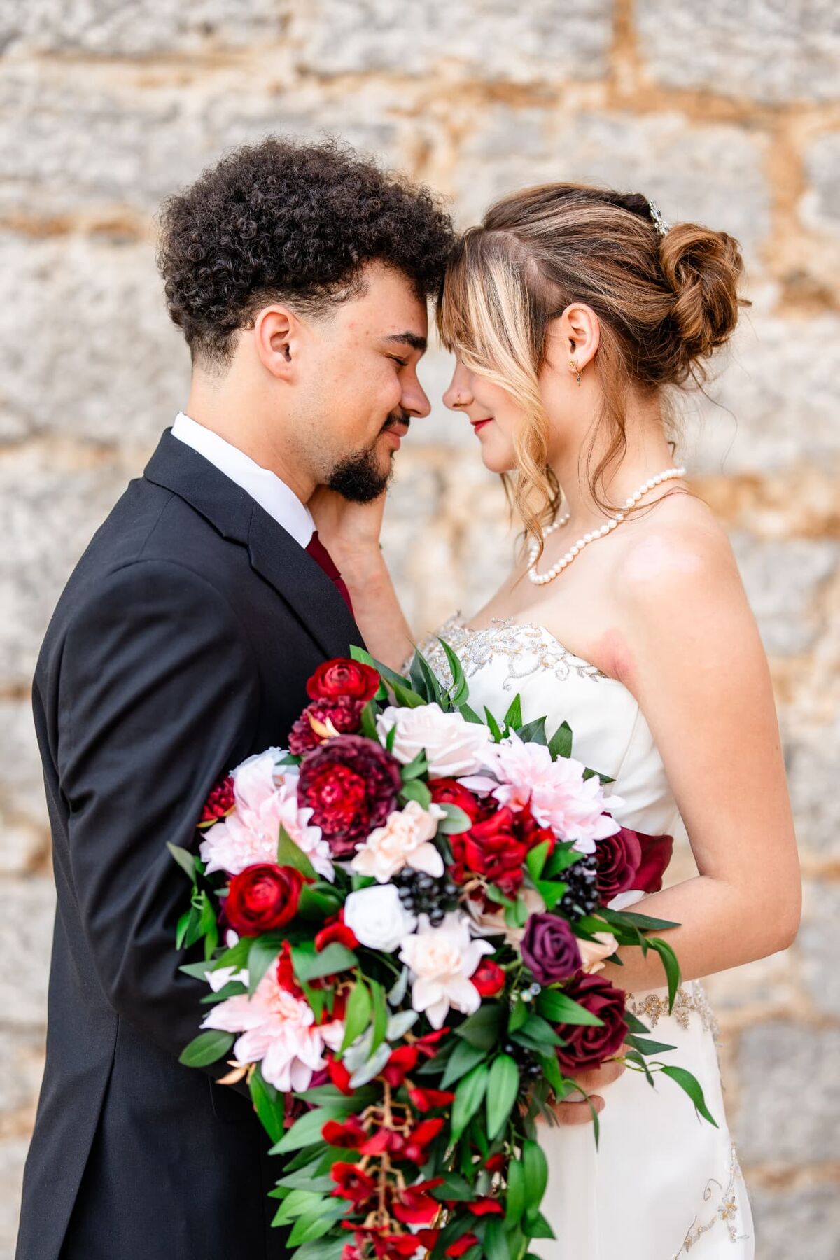 Bride and groom forehead to forehead with a cascading red and blush bouquet