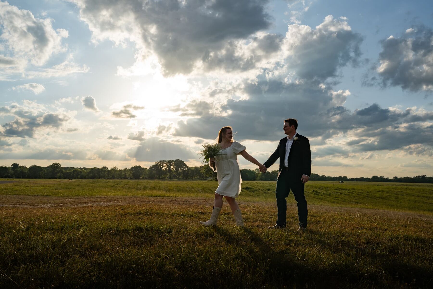 Bride and groom holding hands in an open field at golden hour sunburst